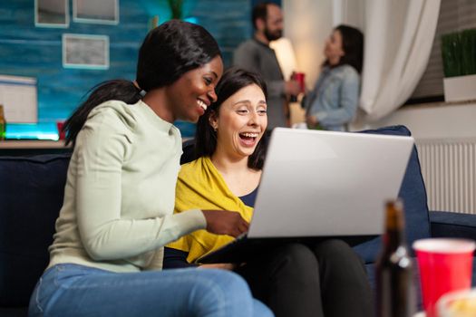 Two multiethnic women sitting on couch watching comedy series on laptop computer hanging out together during night party. In background friends cheering bottles of beer talking and having fun.