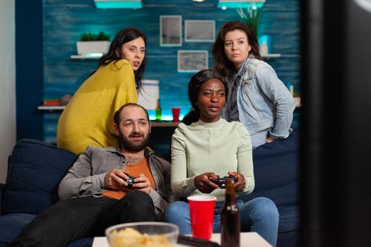 African woman and multi ethnic friends playing video games late in the evening sitting on couch, using wireless controller,socialising. Group of people having fun.