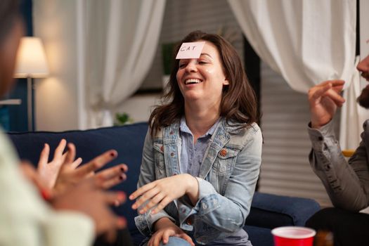 Multiethnic friends having attaching sticky notes on forehead while playing guess who game. Mixed race people having fun, laughing together while sitting on sofa in living room late at night.