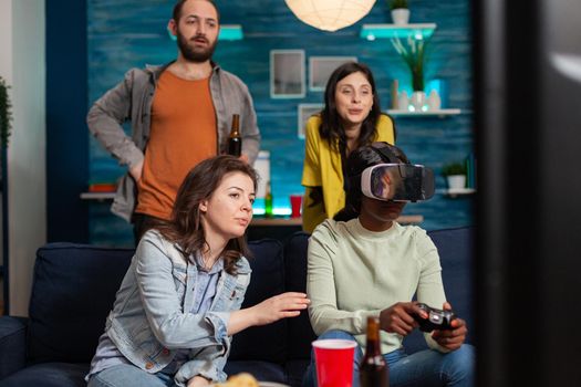 Multi ethnic friends socialising playing video games experiencing virtual reality using headset. Mixed race group of people hanging out together having fun late at night in living room.