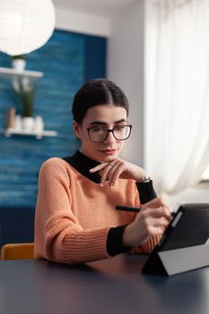 Student artist holding graphic pen in hands drawing illustration sketches on screen of digital tablet computer. Focused woman with glasses sitting at desk table in living room looking at sketch