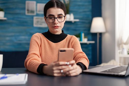 Young woman consulting with her friend using modern phone while sitting at desk table in living room. Student browsing lifestyle information on internet during coronavirus quarantine