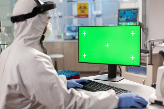 Scientist man wearing protection suit typing on computer with green mockup. Team of microbiologists doing vaccine research writing on device with chroma key, isolated display.