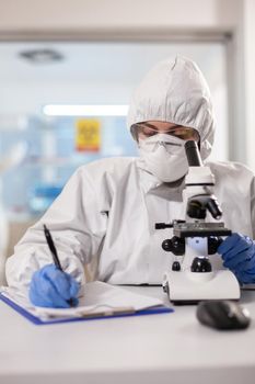 Doctor writing notes dressed in ppe suit and visor mask using microscope. Chemist in coverall working with various bacteria, tissue blood samples for antibiotics research.