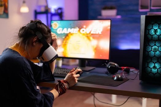 Online player losing shooter competition wearing virtual reality headphones. Defeated player using professional controller for championship sitting on gaming chair late at night in living room