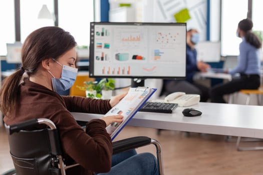 Immobilized business woman with protective mask working in new normal business financial company typing on pc, checking reports, analysing data looking at desktop sitting in wheelchair.
