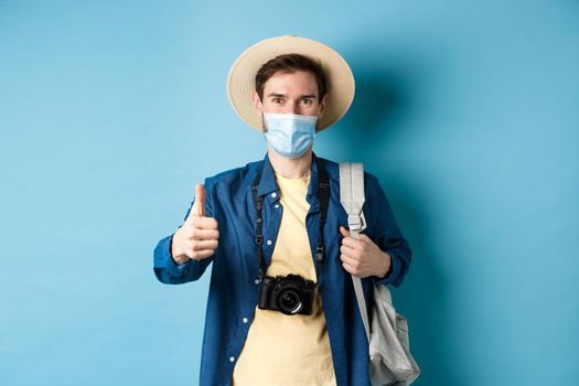 Covid-19 and summer holidays concept. Smiling guy travelling in medical mask and straw hat, backpacking and showing thumbs up, approve and praise travel agency, blue background.