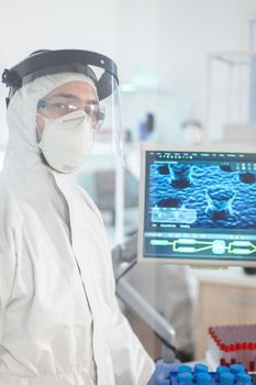 Microbiologist in ppe suit standing in laboratory looking at camera behind the glass wall in equipped lab. Doctor examining virus evolution using high tech and chemistry tools for vaccine development.