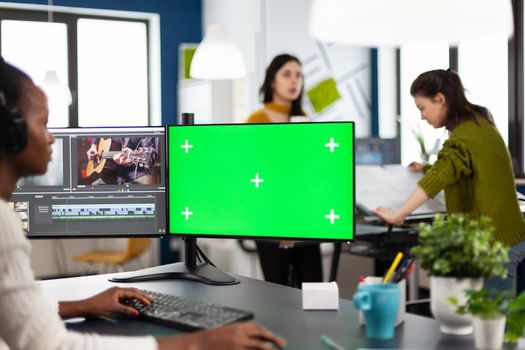 Black woman videographer employee with headphones using computer with green screen, chroma key mockup isolated display sitting in video production studio. African editor processing film montage