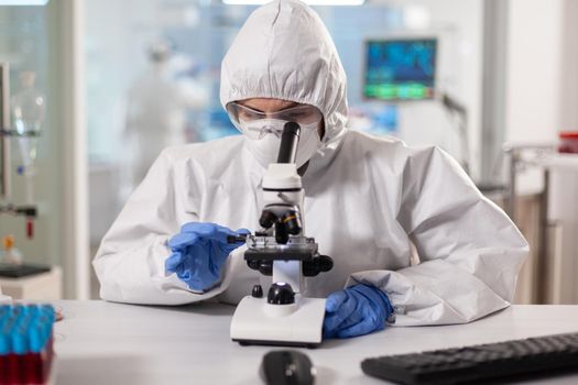 Scientist in ppe suit making adjustments and looking through laboratory microscope. Chemist in coverall working with various bacteria, tissue blood samples for antibiotics research.