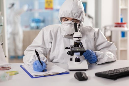 Medicine scientist dressed in ppe suit taking notes during vaccine research using microscope. Chemist in coverall working with various bacteria, tissue blood samples for antibiotics research.