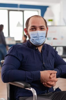 Employee with disabilities, invalid, handicapped paralized with face mask against coronavirus looking at camera sitting immobilized in wheelchair in business office room, working at financial project.