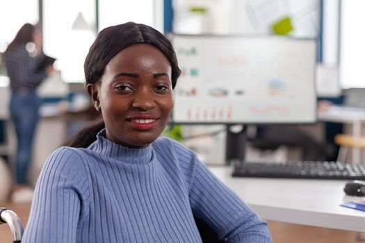 Happy african businesswoman with disabilities looking at camera smiling in immobilized paralysed in wheelchair in business economic office, working on pc with data prosessing financial information.