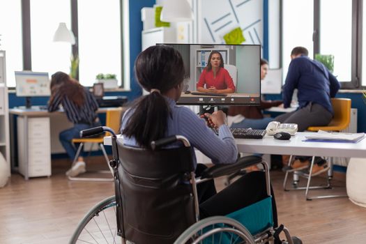 African disabled handicapped businesswoman sitting immobilized in wheelchair talking with remote partner on video call from startup business office. Black paralysed employee discussing project online.