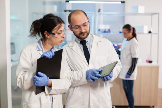 Group of medical researchers discussing about vaccine development, standing in equipped lab pointing on tablet and taking notes. Doctors examining virus evolution using high tech researching diagnosis.