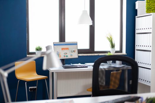 Monitor with financial graphs charts statisctis in corporate office workplace with nobody in it. Empty stylish space of start up workspace. Comfortable chair to sit during long work hours.