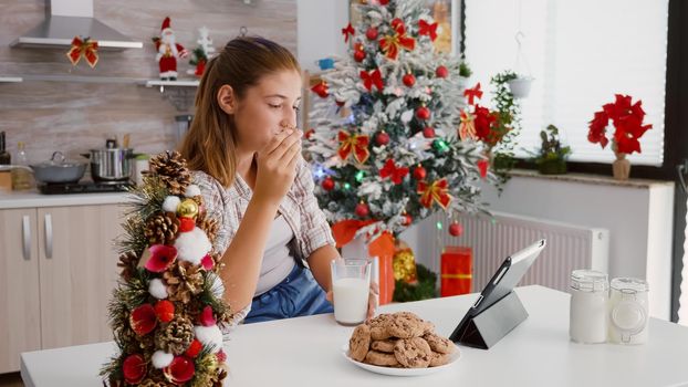 Happy child sitting at table in decorated kitchen watching xmas online video on tablet computer eating baked delicious cookies drinking milk celebrating christmas season. Girl enjoying winter holiday