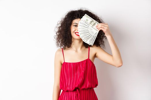 Excited woman in red dress winning money, showing dollar bills and smiling happy, standing on white background.