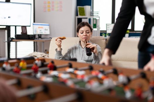 Woman sitting at foosball table at office after work with colleagues eating pizza drinking having fun. Caucasian person watch playing soccer with friends coworkers for entertainment
