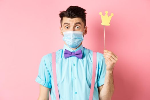 Covid, pandemic and quarantine concept. Cute and silly guy in medical mask looking surprised, holding small crown for party, standing over pink background.