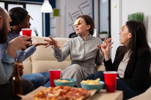 Diverse group of friends play guess who game after work while eating pizza chips and drinking beer. Multi ethnic colleagues enjoy fun cheerful activity at office with food on table