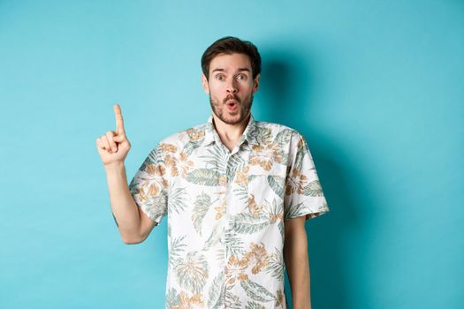 Surprised caucasian tourist in hawaiian shirt, pointing finger up and say wow, checking out promotion, standing on blue background.