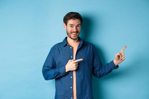 Cheerful caucasian man with beard pointing fingers aside at empty space and smiling at camera, showing advertisement, blue background.