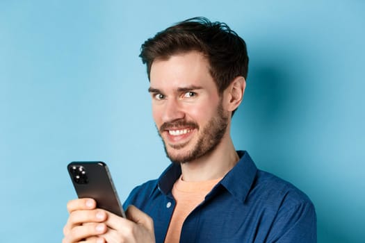 Close-up of cheerful bearded man using smartphone, smiling confident at camera, standing on blue background. Copy space