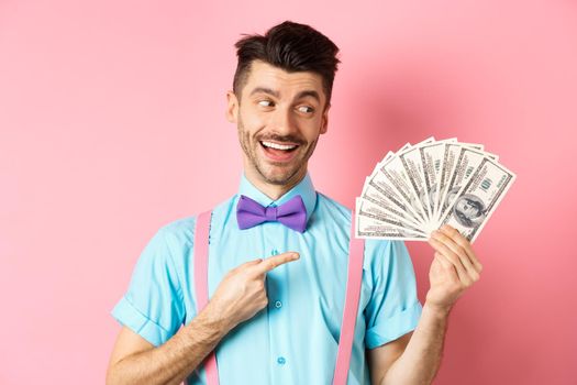 Rich handsome man in festive outfit pointing at money, holding dollars and looking happy, standing on pink background. Copy space