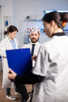 Researcher neurologist doctor asking man's illness symptoms looking at clipboard before brain scan with brainwave scanning headset. Scientist analyzing health status, nervous system, tomography scan.