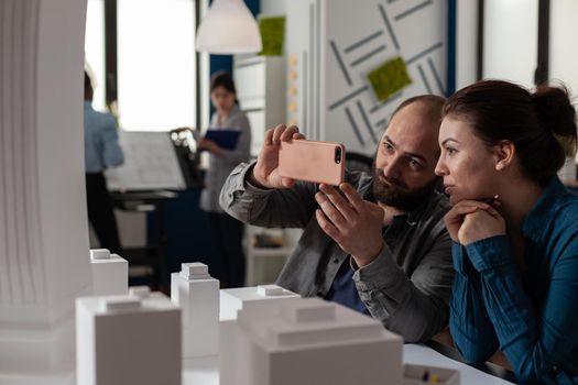 Professional architect colleagues work on smartphone sitting at desk while looking at building model maquette. Engineering workmates talking about construction industry project