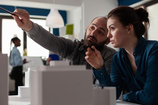 Working architect colleagues discussing layout project to design building model maquette. Professional caucasian workmates pointing at modern construction plan and blueprint