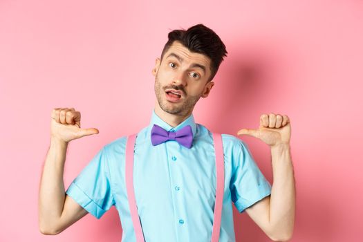 Sassy and confident young man in suspenders and bow-tie, pointing at himself and looking like professional, standing over pink background.