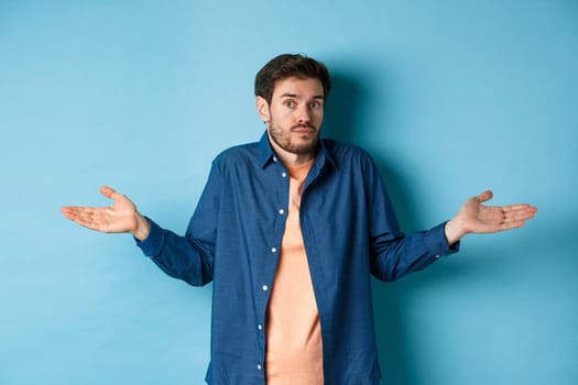 Confused young man with beard, shrugging shoulders and looking clueless at camera, know nothing, standing on blue background.