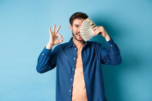 Cheerful young man showing OK sign and cover half of face with dollars, standing on blue background.