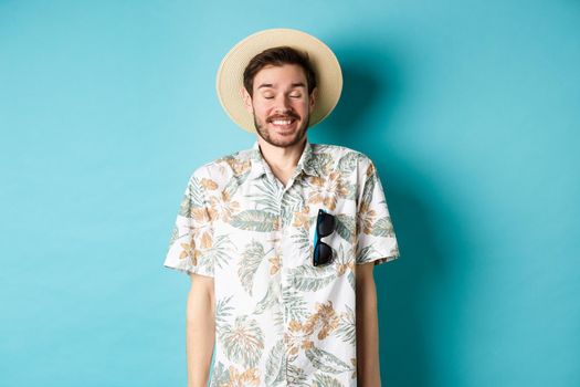 Happy tourist enjoying summer holiday, wearing straw hat and hawaiian shirt, standing joyful on blue background.