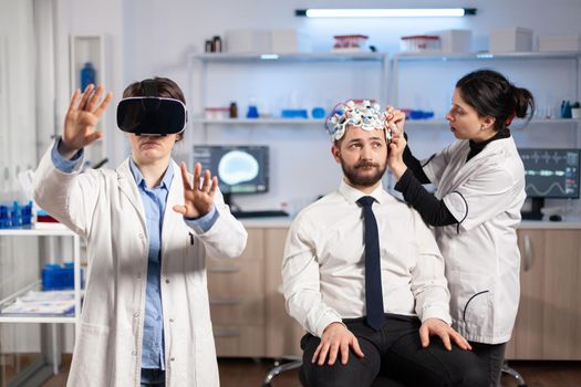 Neuroscientist developing diagnosis wearing virtual reality headset in medicine laboratory, assistant adjusting examining sensors on patient sitting on chair. Medicine lab with modern technology.