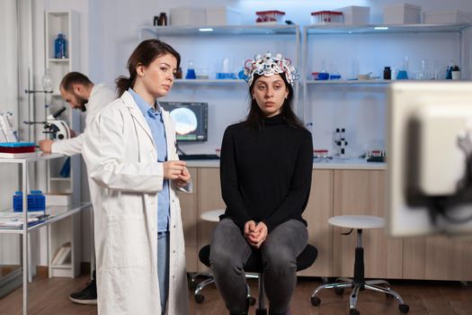 Stressed patient sitting on neurological chair with eeg headset, researcher examining health status writing on tablet. Medical scientist looking through microscope in background developing treatment.