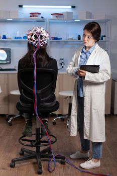 Woman patient wearing performant eeg headset scanning brain electrical activity in neurological research laboratory while medical researcher adjusting it, examining nervous system typing on tablet.