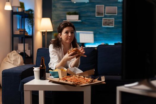 Woman sitting on sofa eating tasty delicious burger watching documentary movie on television during fast-food home delivered in living room. Caucasian female enjoying takeaway junk-food order