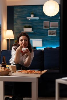 Caucasian female watching entertainment movie on television during takeaway food delivery in evening. Woman eating tasty delicious chinese-food relaxing on sofa enjoying unhealthy night
