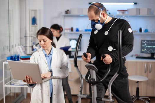 Man athlete running on cross trainer with electrodes attached to his body and mask. Physician usinglaptop and controls EKG data showing on laboratory monitors, discussing with patient.