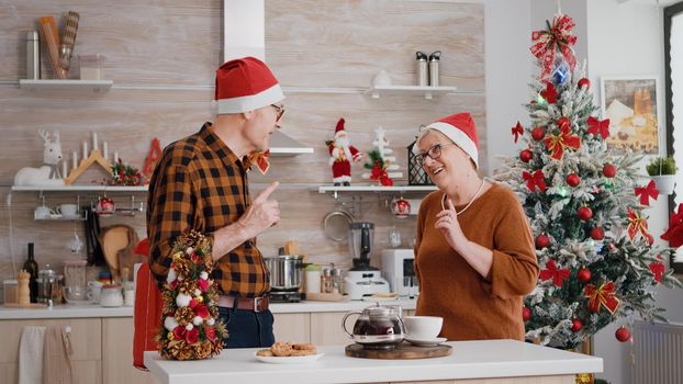 Happy grandparents couple family sharing wrapper gift with ribbon on it enjoying christmastime. Family celebrating winter holiday season surprinsing with present in decorated kitchen