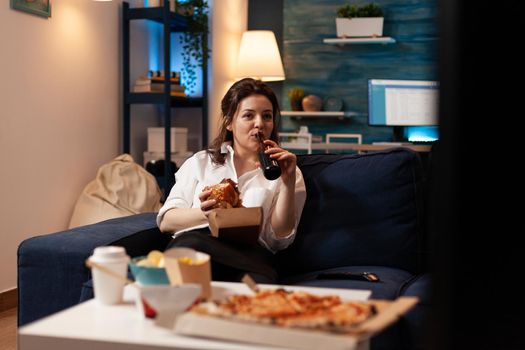 Woman relaxing on couch holding tasty delicious burger while drinking beer watching entertainment movie series on television. Caucasian female enjoying takeaway food home delivered at night