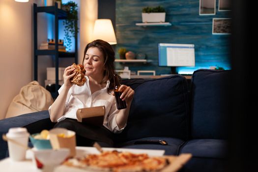 Happy woman eating tasty delicious delivery burger relaxing on sofa watching comedy movie on television in living room. Caucasian female enjoying takeaway fast home delivered in evening