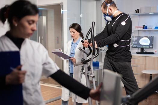 Man athlete running witjh biomechanics attached to his body and mask. Physician looking at notes controls EKG data showing on laboratory monitors, discussing with patient.