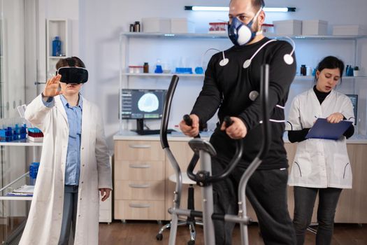 Scientist doctor in sport science laboratory wearing virtual reality goggles while athlete running, with electrodes attached on body monitoring physical endurancewhile ekg scan runs on computer screen.