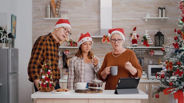 Grandparents with granddaughter greeting remote friends during online videocall meeting in xmas decorated kitchen. Happy family enjoying winter season celebrating christmas