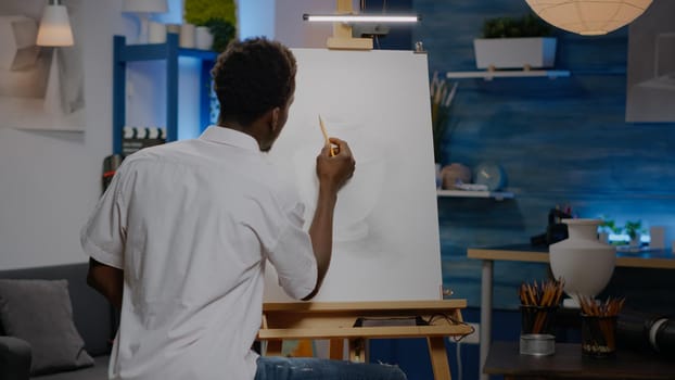 African american young adult using pencil on canvas drawing while looking at white vase for authentic inspiration. Black creative artist creating design for professional masterpiece