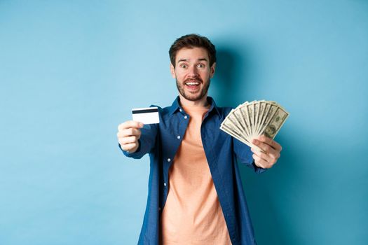 Cheerful caucasian guy extending hands with dollars and plastic credit card, smiling happy at camera, standing on blue background.